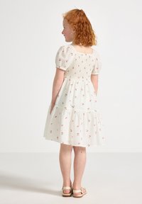 Hvid floral kjole med korte puffærmer, smocked bodice, lagdelt nederdel og røde kirsebærmønstre; parret med lyse sandaler.