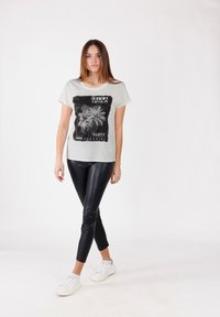 Mujer joven vistiendo una camiseta gráfica blanca de fiesta Ibiza, leggings negros de aspecto cuero y zapatillas blancas, de pie contra un fondo blanco liso.