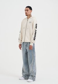 Felpa con zip beige, dotata di tasche frontali e testo nero sulle maniche, abbinata a jeans a gamba larga di un azzurro chiaro e strappati. Sneakers bianche.