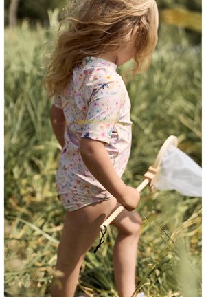 Short-sleeve floral rash guard in pastel colors, featuring a high neck and zip fastening, paired with beige shorts and a wooden net.