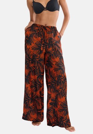 Žena oblečená v čiernom bikinovom topi a voľných tehlovo-oranžových nohaviciach s čiernym tropickým listovým vzorom, ruky v vreckách, bosá na bielom pozadí.