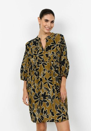 Frau lächelt, trägt ein locker sitzendes olivgrünes Kleid mit schwarzen und beigen Blumenmustern und dreiviertellangen Puffärmeln vor weißem Hintergrund.