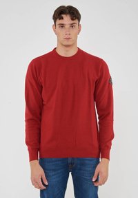 Maglione rosso a maglia con maniche lunghe e scollatura rotonda. Presenta un tessuto texturizzato e una toppa con logo sulla manica sinistra.