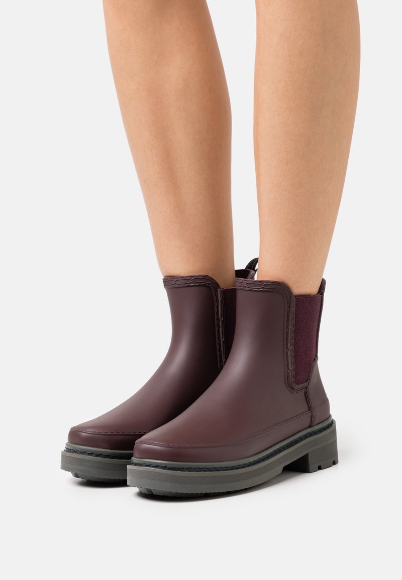 Bottes basses en caoutchouc bordeaux avec panneaux latéraux élastiques, coutures texturées et une semelle épaisse grise. Finition lisse avec des bouts arrondis.