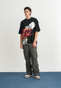 Svart oversized t-shirt med röd "NIKE" grafik och bilder av basket; matchad med olivgröna cargobyxor och neutrala sneakers.