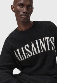Juodas megztinis su balta "ALLSAINTS" tekstu priekyje. Turi kraštuotą apvalią iškirptę ir tekstūruotą medžiagą.