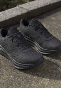 Paire de chaussures de course noires avec dessus en mesh texturé et trois bandes latérales, posées sur le trottoir près d'un bord.