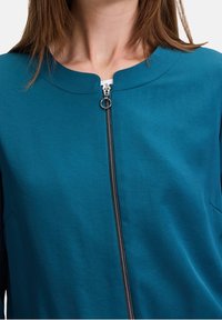 Teal zip-up top featuring a round neckline, smooth fabric texture, and silver zipper detail down the center.