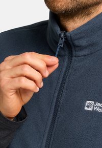 Dunkelblaue Fleecejacke mit hohem Kragen, Reißverschluss und Logo. Eine Hand zieht den Reißverschlussschieber nach, wodurch die weiche Textur und die Nähdetails sichtbar werden.