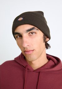 Mørkebrun strikket hue med ribbet tekstur og et lille rundt logo-patch, båret med en maroon hoodie. Nærbillede af ansigtet.