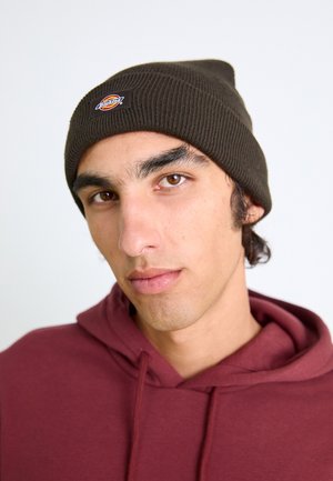 Dunkelbraune gestrickte Mütze mit gerippter Textur und einem kleinen runden Logo-Patch, getragen mit einem kastanienbraunen Hoodie. Nahaufnahme des Gesichts.