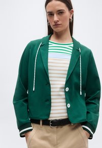 Νεαρή γυναίκα που φοράει πράσινο σακάκι με κουμπιά και λευκές κορδονάκια, ριγέ πουκάμισο, μπεζ παντελόνι και μαύρη ζώνη, με τα χέρια στις τσέπες και ουδέτερη έκφραση.