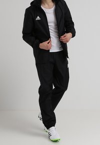 Henkilö, joka on pukeutunut mustaan Adidas-trainingspukuun, valkoiseen T-paitaan ja valkoisiin Adidas-tennareihin, joissa on vihreät pohjat, seisoo tasaisella taustalla.