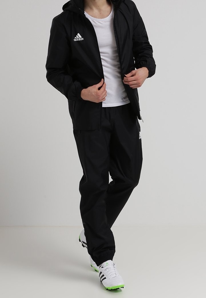 Henkilö, joka on pukeutunut mustaan Adidas-trainingspukuun, valkoiseen T-paitaan ja valkoisiin Adidas-tennareihin, joissa on vihreät pohjat, seisoo tasaisella taustalla.