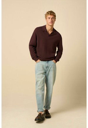 Junger Mann mit Händen in den Taschen, trägt ein dunkelbraunes langärmliges Poloshirt, hellblaue hochgekrempelte Jeans und dunkelbraune Loafers, stehend vor beigem Hintergrund.