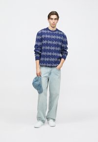 Granatowy sweter z białym wzorem szkieletu, z okrągłym dekoltem i ściągaczami na rękawach, w połączeniu z jasnoniebieskimi dżinsami i jeansową czapką. Białe buty.