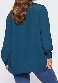 Teal, long-sleeve blouse with a relaxed fit, lightweight material, button accents on the sleeves, and a slightly longer back hem.