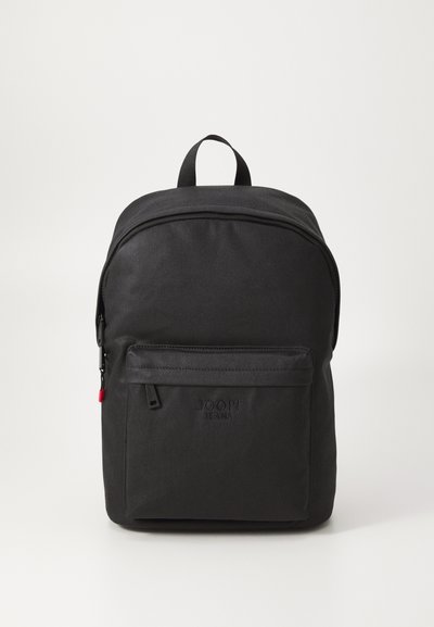 Sac à dos en tissu noir avec une poignée supérieure, fermeture à glissière incurvée, poche avant et détail de logo. Texture lisse et design minimaliste.