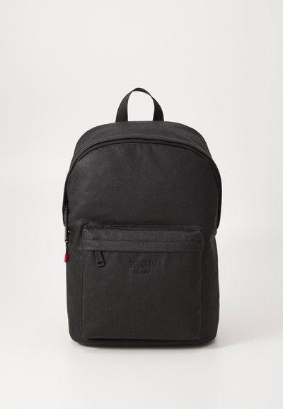 Sac à dos en tissu noir avec une poignée supérieure, fermeture à glissière incurvée, poche avant et détail de logo. Texture lisse et design minimaliste.