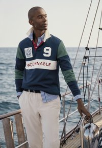 Marineblauwe polo met blokkleurige mouwen in groen en blauw, witte band met de tekst "FACONNABLE", gecombineerd met beige broek en gestreept shirt eronder.
