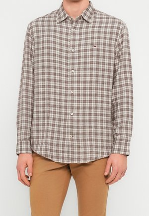 Homme portant une chemise à carreaux beige et marron à manches longues et un pantalon beige, debout devant un fond uni.