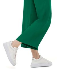Sneakers in pelle bianca con una tomaia testurizzata, caratterizzate da cuciture decorative, una chiusura con lacci e una suola spessa in gomma. Abbinati a pantaloni verdi.