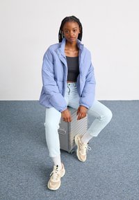 Veste matelassée bleu clair avec un col montant, des poignets élastiques et une fermeture à pressions à l'avant, associée à un jean bleu clair et des baskets multicolores.