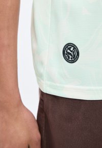 Detailný záber na trup osoby v svetlozelenom športovom drese s čiernym emblémom "Authentic Licensed Football" nad hnedými nohavicami.