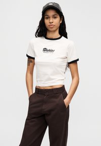 Λευκό κοντό μπλουζάκι με μαύρο στρογγυλό λαιμόκοψη, με το λογότυπο "Dickies Workwear Company". Καφέ ψηλόμεσο παντελόνι. Ομαλή υφή υφάσματος.