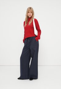 Pull à col roulé rouge côtelé, pantalon large rayé bleu marine ample, et sac à épaule blanc. Des chaussures noires complètent la tenue.