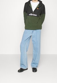 Zielona i czarna polarowa kurtka z kapturem, z zamkiem na pół i logo, zestawiona z jasnoniebieskimi szerokimi jeansami oraz czarnymi butami. Minimalistyczny design.