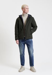 Giacca con cappuccio di un verde scuro, dalla texture liscia, indossata sopra un dolcevita beige chiaro, abbinata a jeans in denim blu e sneakers beige.