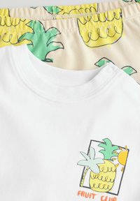 Witte katoenen t-shirt met een kleurrijke ananasafbeelding met groene bladeren en zon, gecombineerd met gele en crèmekleurige shorts met een ananaspatroon.