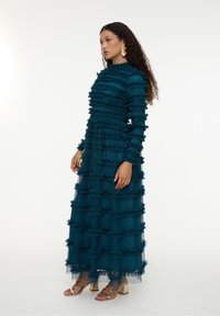 Teal värjätty tiered-kjol med rynkig textur, långa ärmar och hög halsringning, kompletterad med metalliska blockklacksandaler.