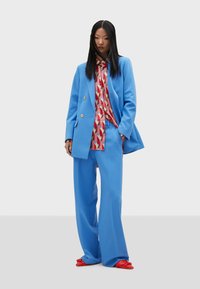 Blue tailored suit with double-breasted blazer and wide-leg trousers. Underneath, a patterned red and pink shirt. Bright red shoes complete the look.