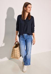 Camicia blu navy a maniche lunghe, jeans a gamba larga azzurri chiari, sneakers bianche e borsa a rete color beige con motivo a griglia nera.