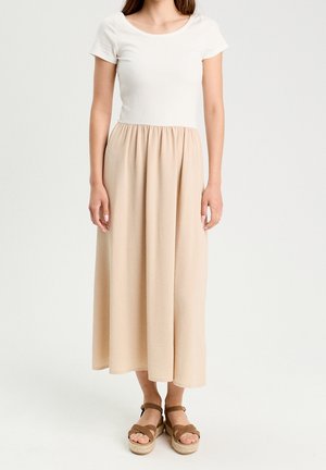 Femme portant un haut blanc à manches courtes et une jupe midi beige taille haute avec des sandales compensées marron, debout devant un fond uni.