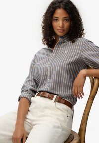 Blå-grå skjorte med gule striber, der har krave og broderet logo; parret med hvide bukser og et brunt bælte.