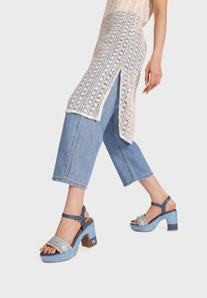 Gambe con jeans azzurri cropped, sandali con tacco alto blu e argento, e una lunga tunica crochet color crema con spacchi laterali.