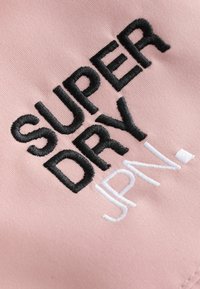 Tela rosa con texto bordado en negro y blanco en relieve que dice "SUPER DRY JPN". Enfoque en costura limpia y textura suave.