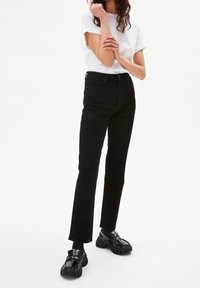 Mujer de pie con jeans rectos negros, mocasines negros gruesos, calcetines negros y una camiseta blanca de manga corta, posando con los brazos cruzados.
