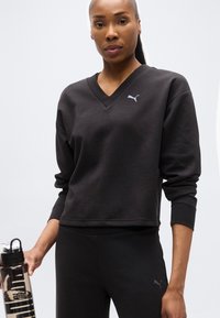 Γυναίκα που φοράει μαύρο φούτερ και παντελόνι Puma, κρατώντας ένα διαφανές μπουκάλι νερού με μαύρο καπάκι μπροστά σε ανοιχτόχρωμο φόντο.