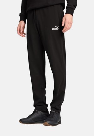 Uomo con pantaloni della tuta neri Puma e sneakers nere, in piedi davanti a uno sfondo chiaro e semplice.