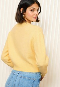 Pull en tricot jaune avec un col rond et un ourlet côtelé. Le modèle porte un jean taille haute bleu, mettant en valeur la texture et la couleur du pull.