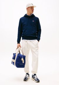 Marine hoodie met trekkoorden, crèmekleurige sweatpants en blauwe sporttas met crèmekleurige banden. Sneakers met marineblauwe en witte accenten.