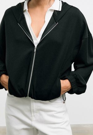 Personne portant un sweat à capuche noir zippé avec des poignets élastiques sur une chemise blanche à col et un pantalon blanc, les mains dans les poches du sweat.