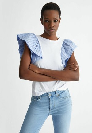 Maglietta bianca con maniche a volant a righe blu, abbinata a jeans azzurri. La maglietta presenta un colletto rotondo e una vestibilità casual.