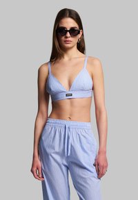 Sininen ja valkoinen raidallinen bralette, jossa on kolmiomaiset kupit, kapeat hihnat ja logo. Yhdistetty yhteensopiviin löysiä raidallisiin housuihin.