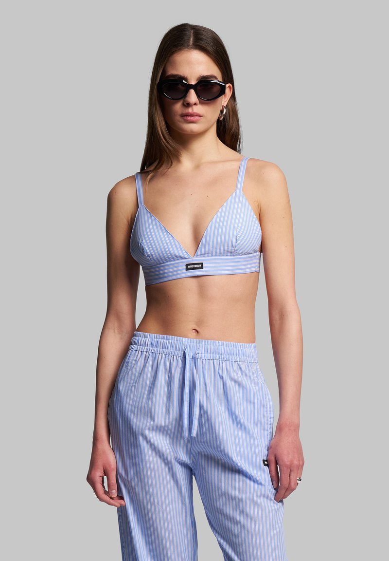 Sininen ja valkoinen raidallinen bralette, jossa on kolmiomaiset kupit, kapeat hihnat ja logo. Yhdistetty yhteensopiviin löysiä raidallisiin housuihin.
