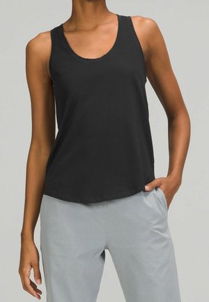 Osoba nosząca czarny bezrękawnik z okrągłym dekoltem i jasnoszare spodnie, z ręką w kieszeni, na jednolitym szarym tle.